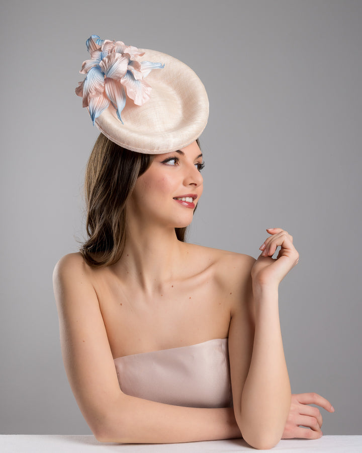 The female model is posing, wearing Marion,the light pink oval high based fascinator decorated with custom made pink and blue shaded flowers. Designed and made by Merve Bayindir Millinery.
