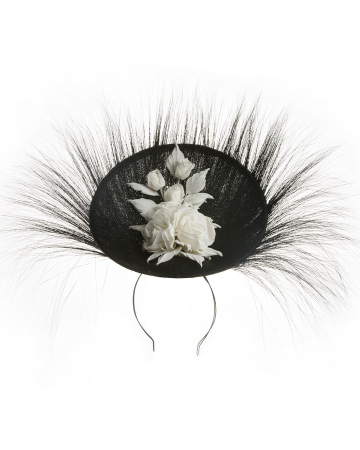 A dramatic, sculptural fascinator featuring a sleek black sinamay disc base. From the edges, long, fine black quills radiate outward in a striking sunburst effect, creating movement and sharp elegance. At the center sits a cluster of handcrafted ivory roses and buds with matching leaves, providing a contrast against the dark base. Designed and made by Merve Bayindir Millinery.

