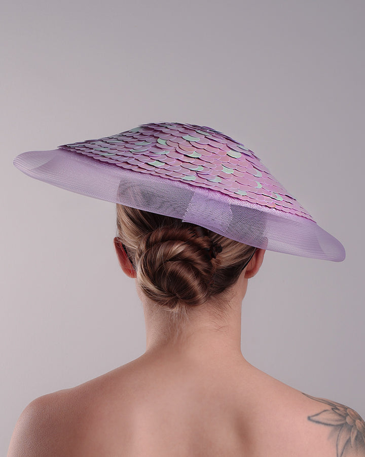 Model is showcasing the back of a circle shaped lilac saucer hat extended with crinoline mesh, fully embroideried with lilac and green sequins.