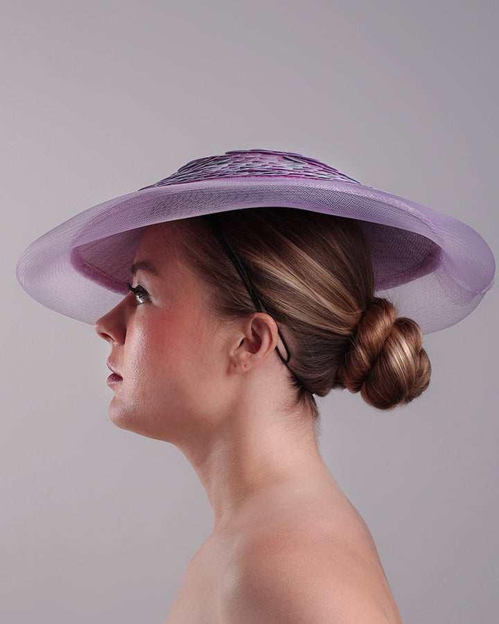 Model is showcasing the left side of a circle shaped lilac saucer hat extended with crinoline mesh, fully embroideried with lilac and green sequins.