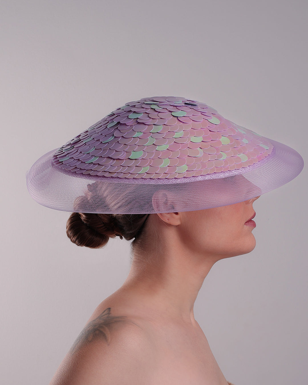 Model is showcasing the right side of a circle shaped lilac saucer hat extended with crinoline mesh, fully embroideried with lilac and green sequins.