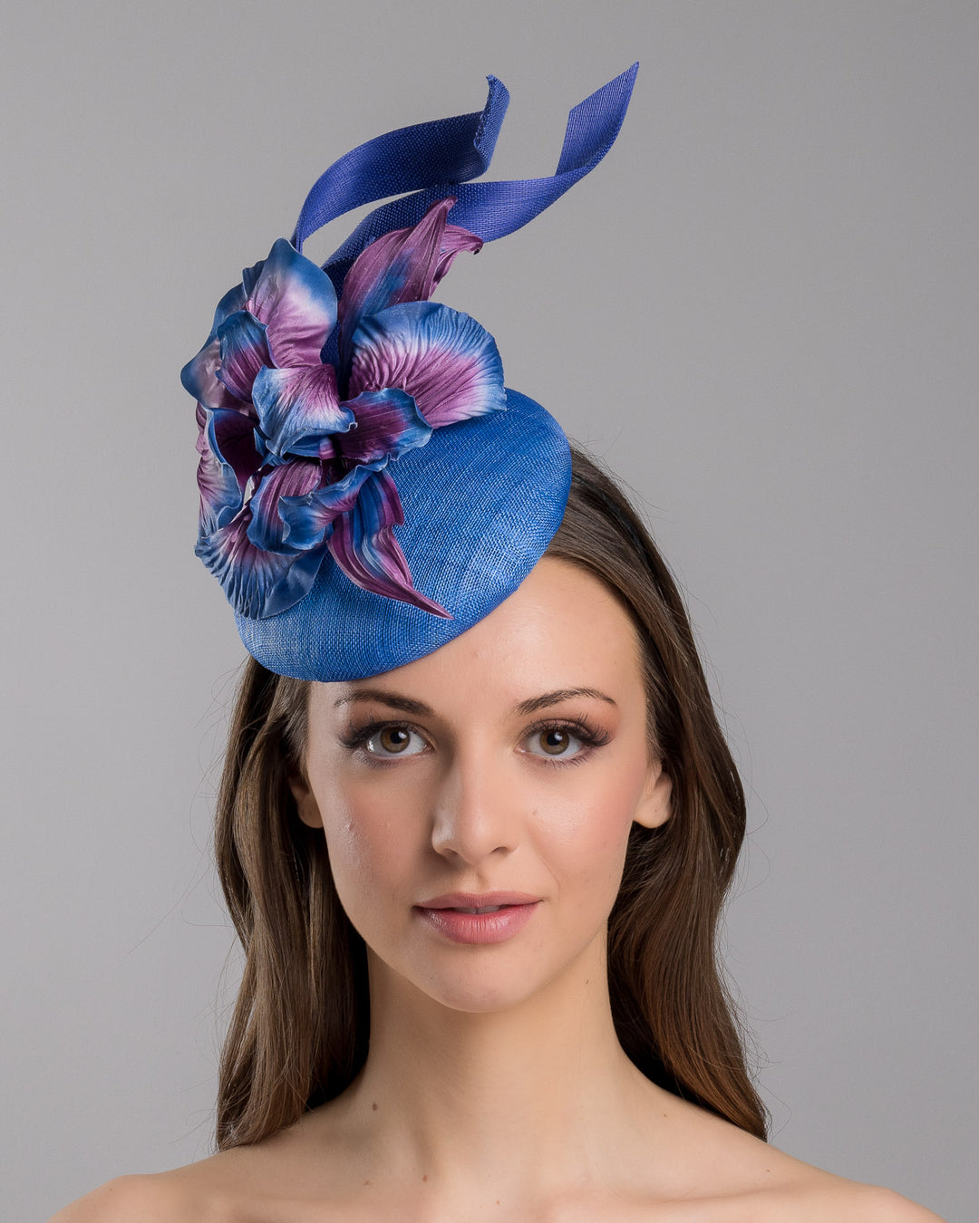 The model is showcasing the front side close up of the deep blue button shaped fascinator decorated with colourful irises and two high buntal swirls. The flower is detailed in transitioning colour from deep blue to purple on iris petals. Designed and made by Merve Bayindir Millinery.
