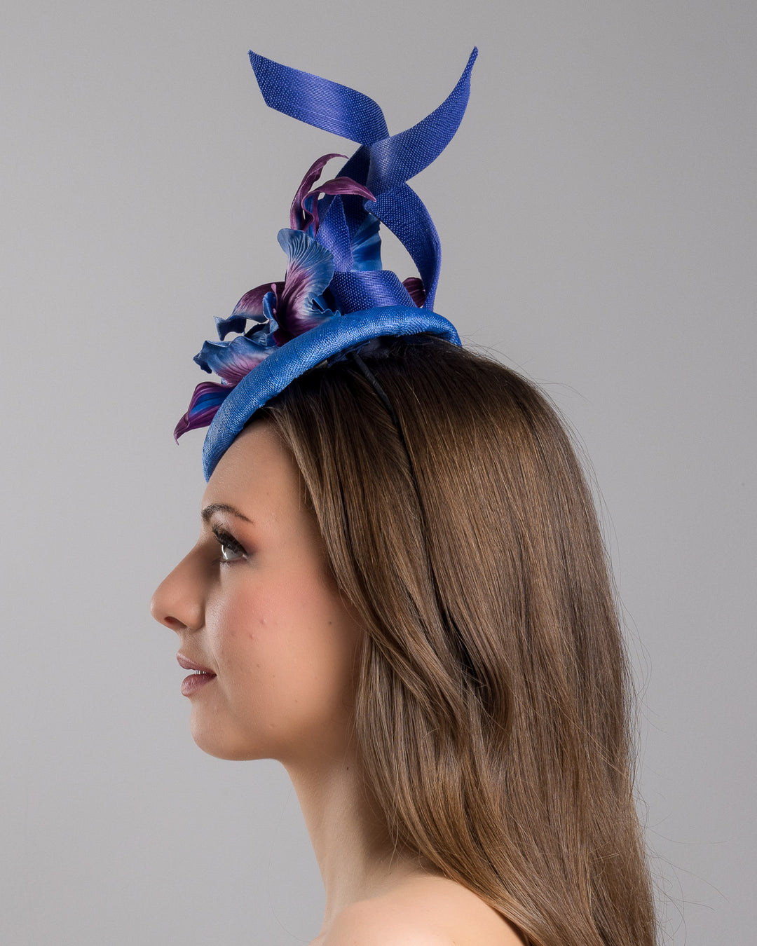 The model is showcasing the left side close up of the deep blue button shaped fascinator decorated with colourful irises and two high buntal swirls. The flower is detailed in transitioning colour from deep blue to purple on iris petals. Designed and made by Merve Bayindir Millinery.

