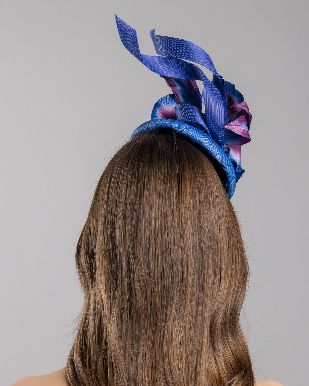 The model is showcasing the back side close up of the deep blue button shaped fascinator decorated with colourful irises and two high buntal swirls. The flower is detailed in transitioning colour from deep blue to purple on iris petals. Designed and made by Merve Bayindir Millinery.
