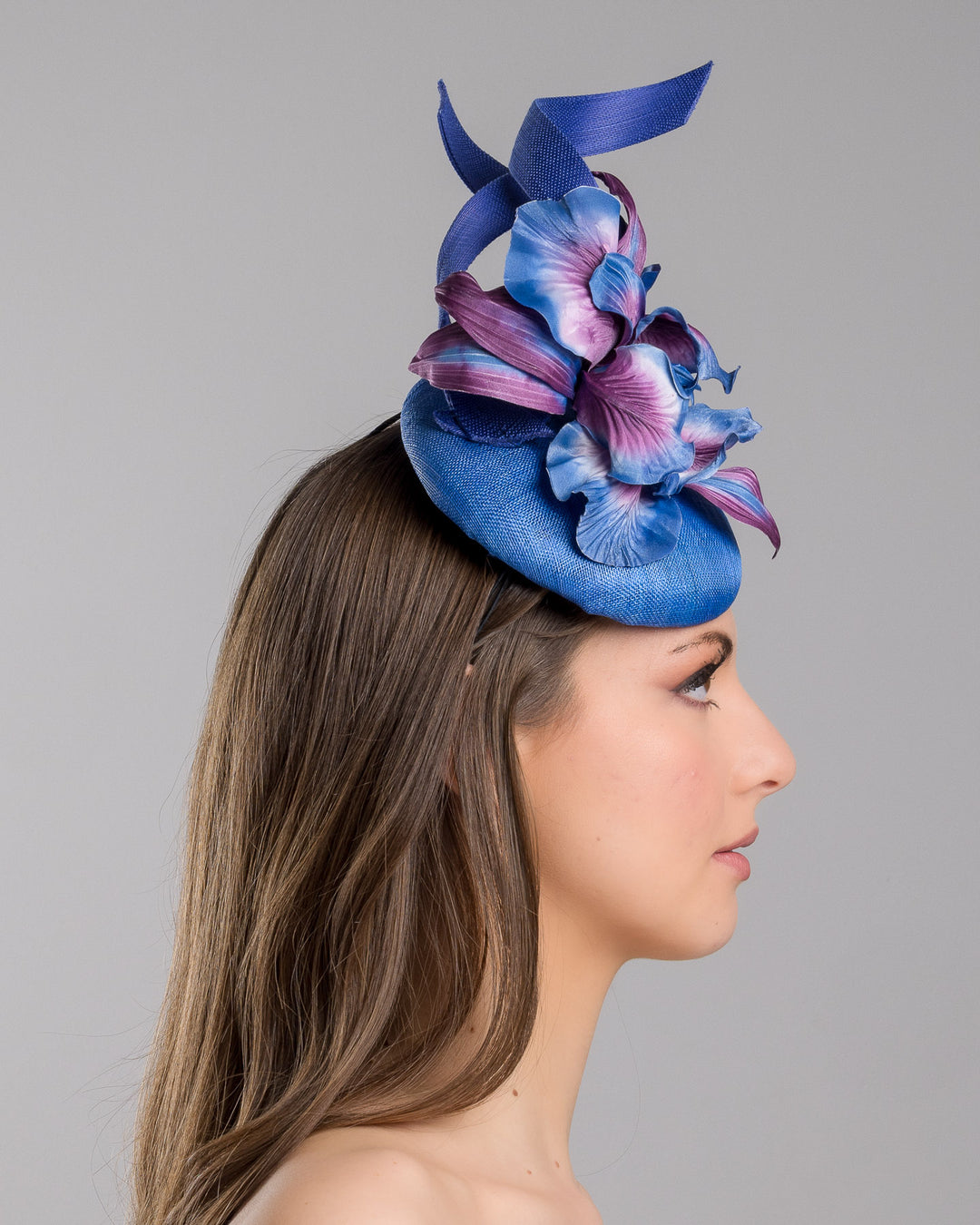 The model is showcasing the right side close up of the deep blue button shaped fascinator decorated with colourful irises and two high buntal swirls. The flower is detailed in transitioning colour from deep blue to purple on iris petals. Designed and made by Merve Bayindir Millinery.
