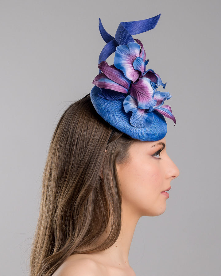 The model is showcasing the right side close up of the deep blue button shaped fascinator decorated with colourful irises and two high buntal swirls. The flower is detailed in transitioning colour from deep blue to purple on iris petals. Designed and made by Merve Bayindir Millinery.
