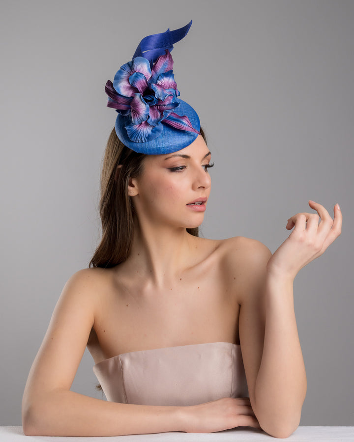 The female model is posing, wearing Max,  the deep blue button shaped fascinator decorated with colourful irises and two high buntal swirls. The flower is detailed in transitioning colour from deep blue to purple on iris petals. Designed and made by Merve Bayindir Millinery.
