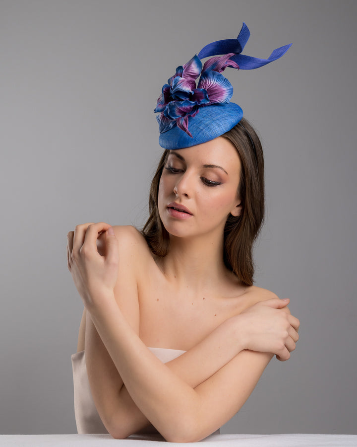 The female model is posing, wearing Max,  the deep blue button shaped fascinator decorated with colourful irises and two high buntal swirls. The flower is detailed in transitioning colour from deep blue to purple on iris petals. Designed and made by Merve Bayindir Millinery.
