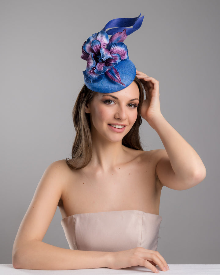 The female model is posing, wearing Max,  the deep blue button shaped fascinator decorated with colourful irises and two high buntal swirls. The flower is detailed in transitioning colour from deep blue to purple on iris petals. Designed and made by Merve Bayindir Millinery.
