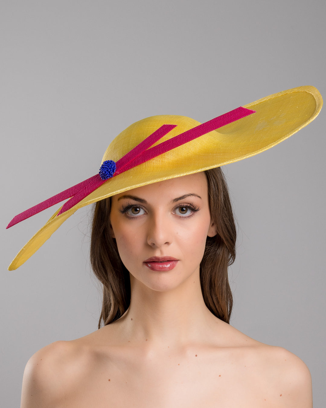 Model is showcasing the front close  up of wide brim asymmetric yellow hat tilted to the left. Decorated with two pink buntal stripes crossing the hat as well as each other, embellished the centre of straight ribbon is a dark blue bead bead cluster. Designed and made by Merve Bayindir millinery.