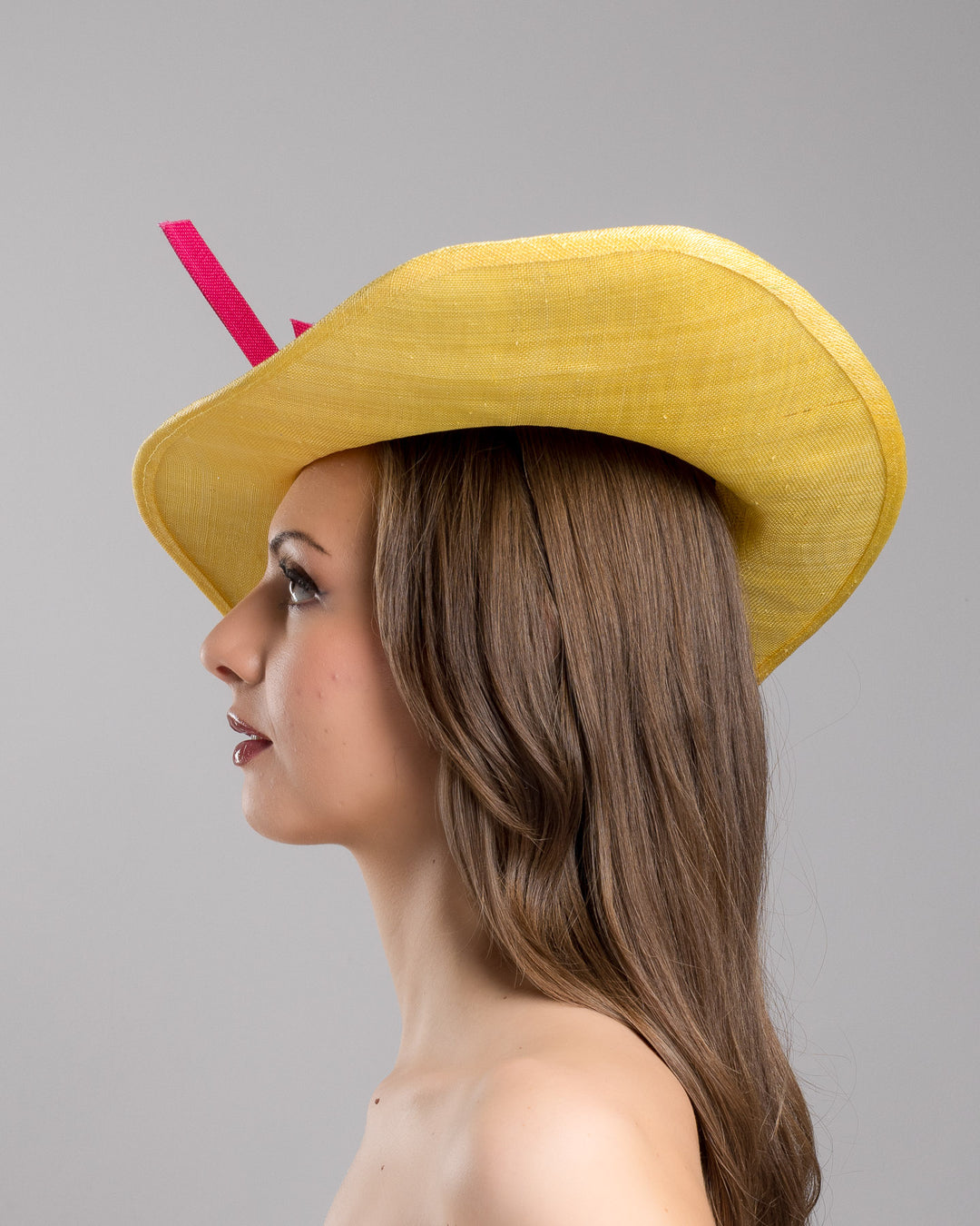 Model is showcasing the left side close  up of wide brim asymmetric yellow hat tilted to the left. Decorated with two pink buntal stripes crossing the hat as well as each other, embellished the centre of straight ribbon is a dark blue bead bead cluster. Designed and made by Merve Bayindir millinery.