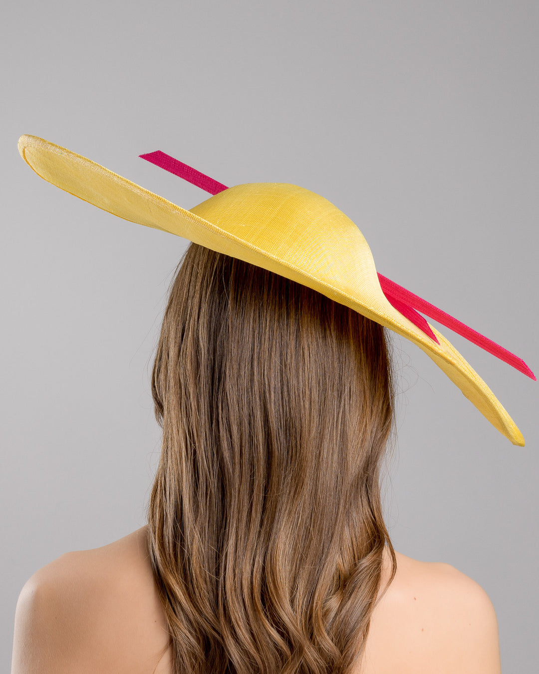 Model is showcasing the back side close  up of wide brim asymmetric yellow hat tilted to the left. Decorated with two pink buntal stripes crossing the hat as well as each other, embellished the centre of straight ribbon is a dark blue bead bead cluster. Designed and made by Merve Bayindir millinery.