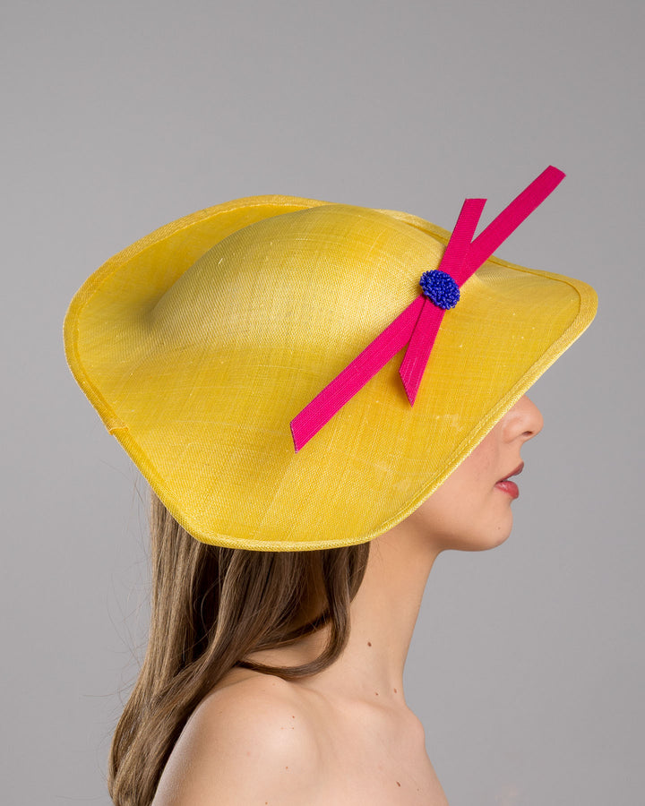 Model is showcasing the right side close  up of wide brim asymmetric yellow hat tilted to the left. Decorated with two pink buntal stripes crossing the hat as well as each other, embellished the centre of straight ribbon is a dark blue bead bead cluster. Designed and made by Merve Bayindir millinery.