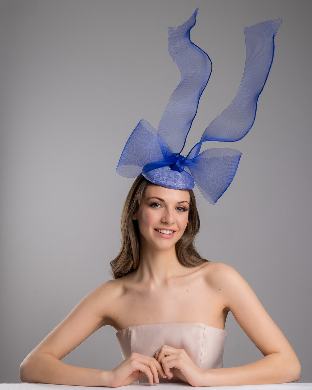 Model is wearing  Royal blue button shaped fascinator decorated with a large crinoline bow and flying crinoline ribbons. Made by Merve Bayindir Millinery.