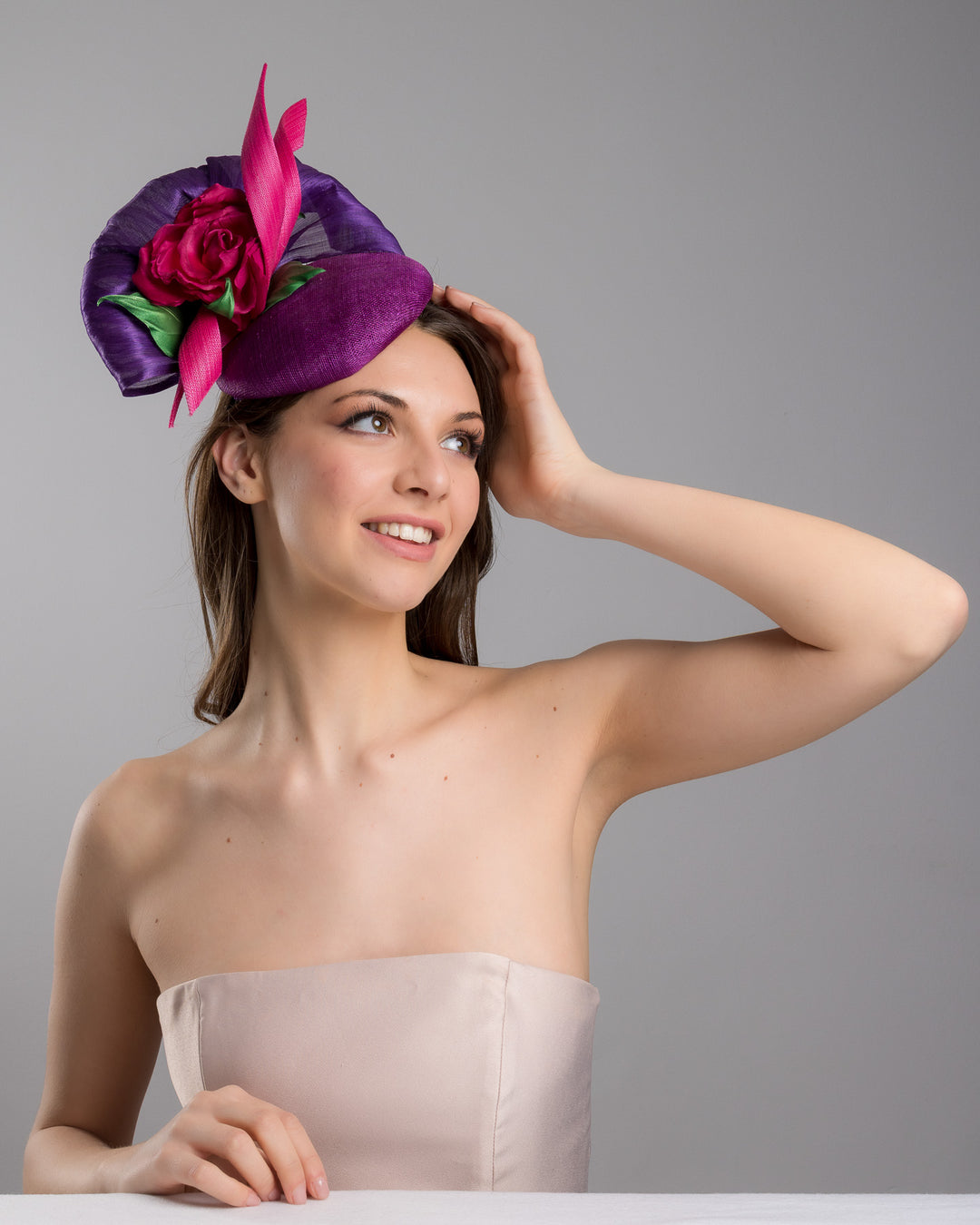 The female model is posing, wearing Tanner, the purple sinamay button shaped fascinator decorated with curved silk abaca fabric curving like a sea shell, two buntal swirls and a large pink silk rose, exemplifying the harmony of different shades of color. Designed and made by Merve Bayindir Millinery.
