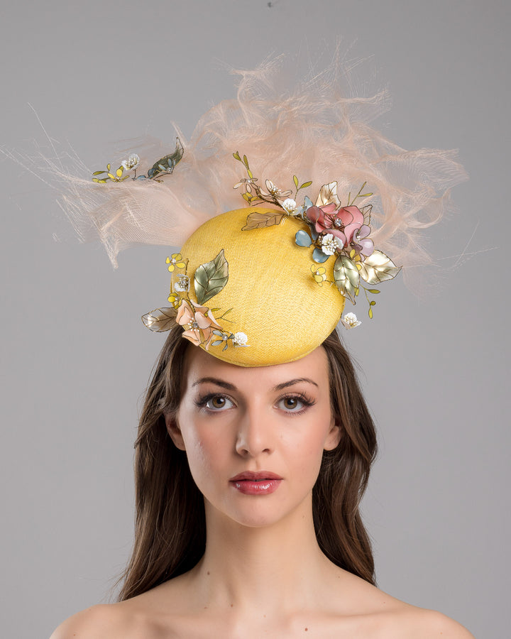 Model is showcasing the front of the double button yellow fascinator decorated with multi colour  wired resin flowers and peach crinoline clouds. Made by Merve Bayindir Millinery.
