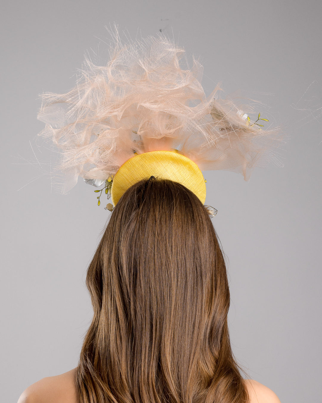 Model is showcasing the back of the double button yellow fascinator decorated with multi colour  wired resin flowers and peach crinoline clouds. Made by Merve Bayindir Millinery.
