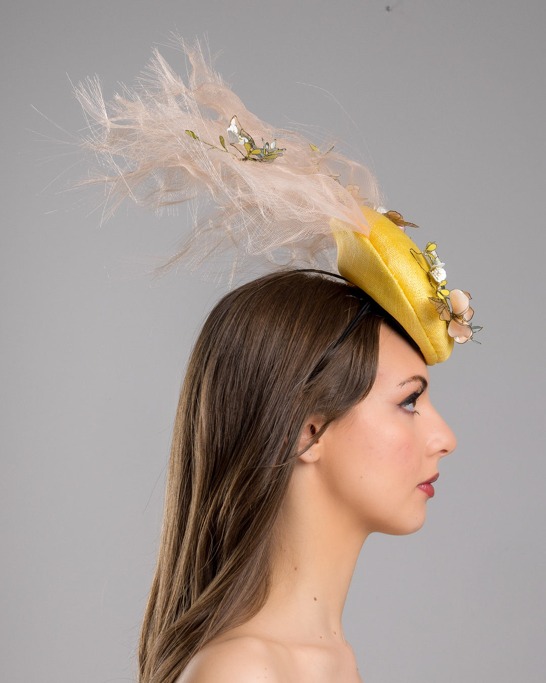 Model is showcasing the right side of the double button yellow fascinator decorated with multi colour  wired resin flowers and peach crinoline clouds. Made by Merve Bayindir Millinery.