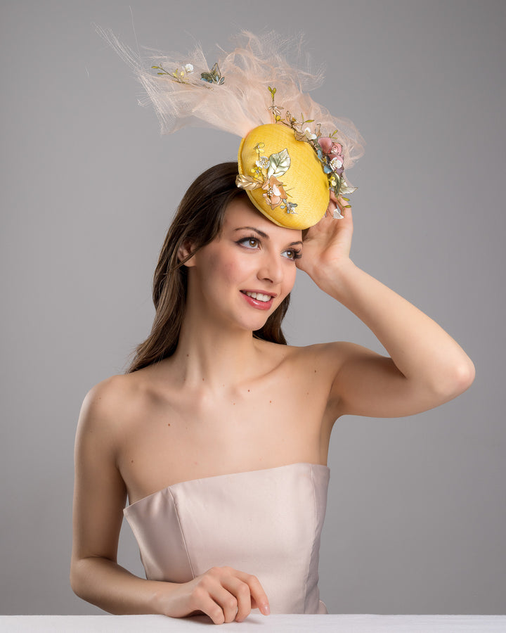 Model is posing with Merve Bayindir double button yellow fascinator decorated with multi colour  wired resin flowers and peach crinoline clouds. Made by Merve Bayindir Millinery.