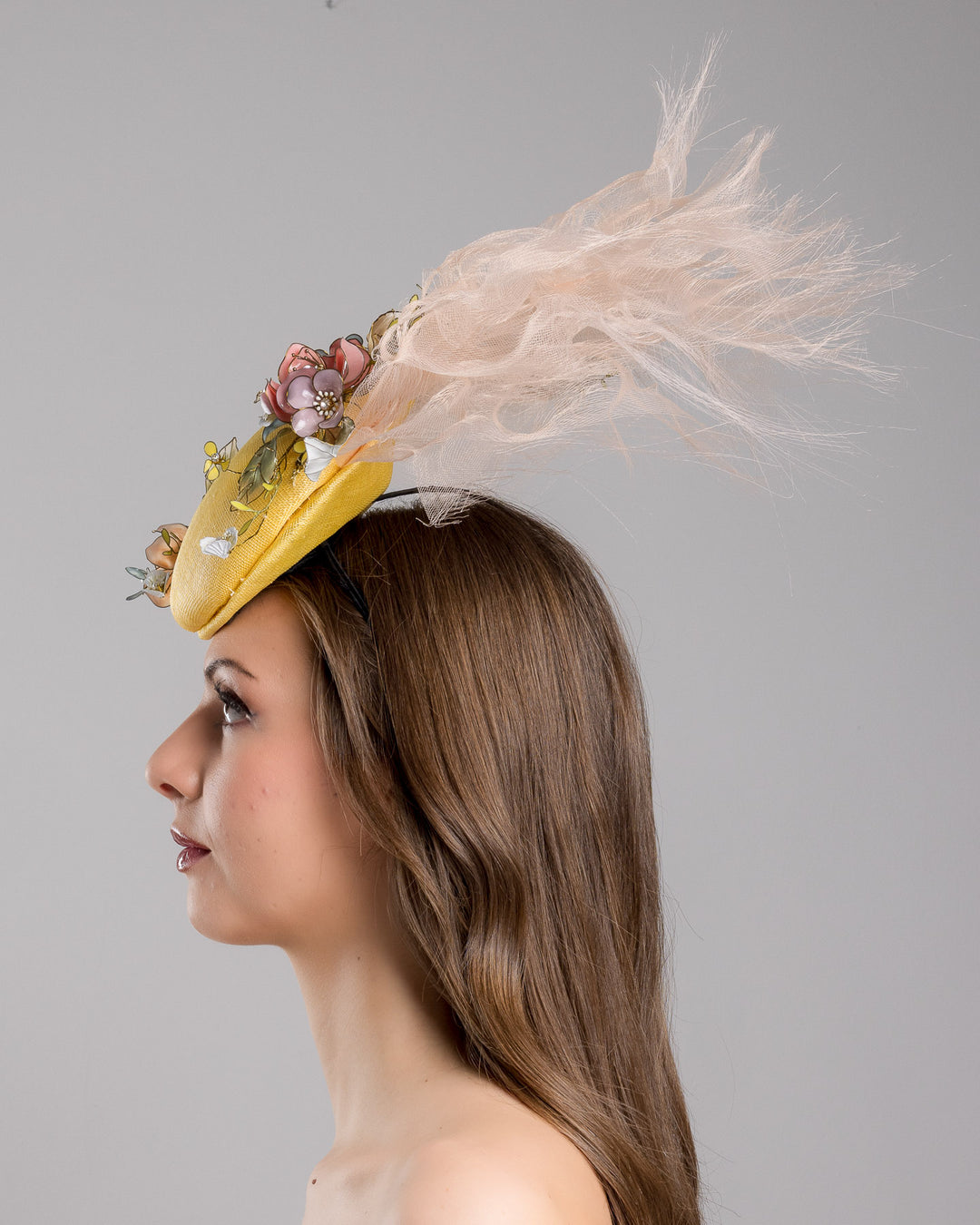 Model is showcasing the left side of the double button yellow fascinator decorated with multi colour  wired resin flowers and peach crinoline clouds. Made by Merve Bayindir Millinery.