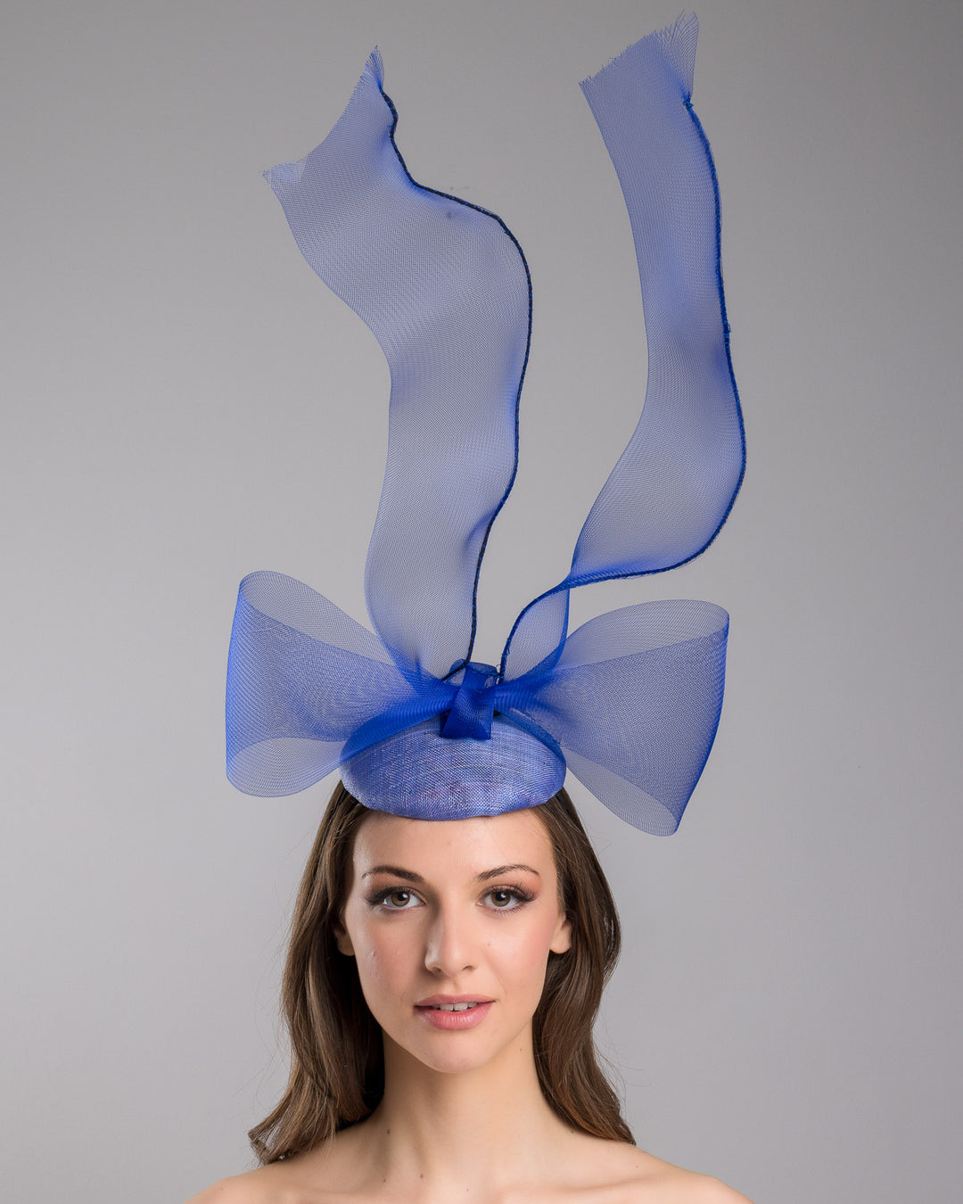 Model is showcasing the front of the Royal blue button shaped fascinator decorated with a large crinoline bow and flying crinoline ribbons. Made by Merve Bayindir Millinery.