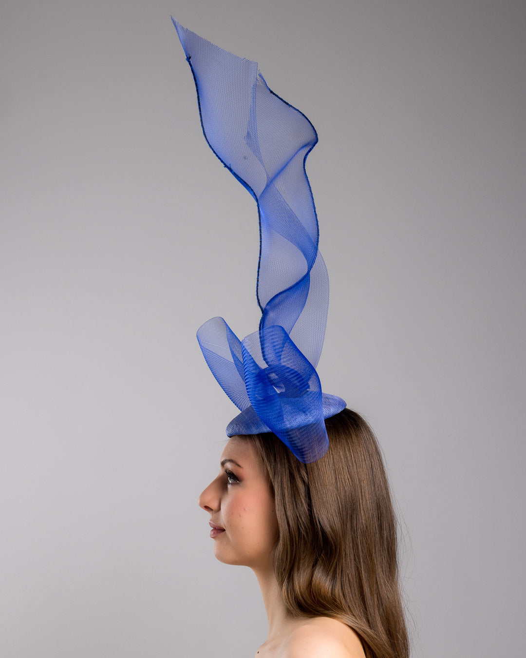 Model is showcasing the left side of the Royal blue button shaped fascinator decorated with a large crinoline bow and flying crinoline ribbons. Made by Merve Bayindir Millinery.