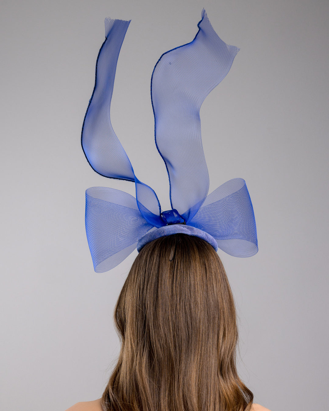 Model is showcasing the back of the Royal blue button shaped fascinator decorated with a large crinoline bow and flying crinoline ribbons. Made by Merve Bayindir Millinery.