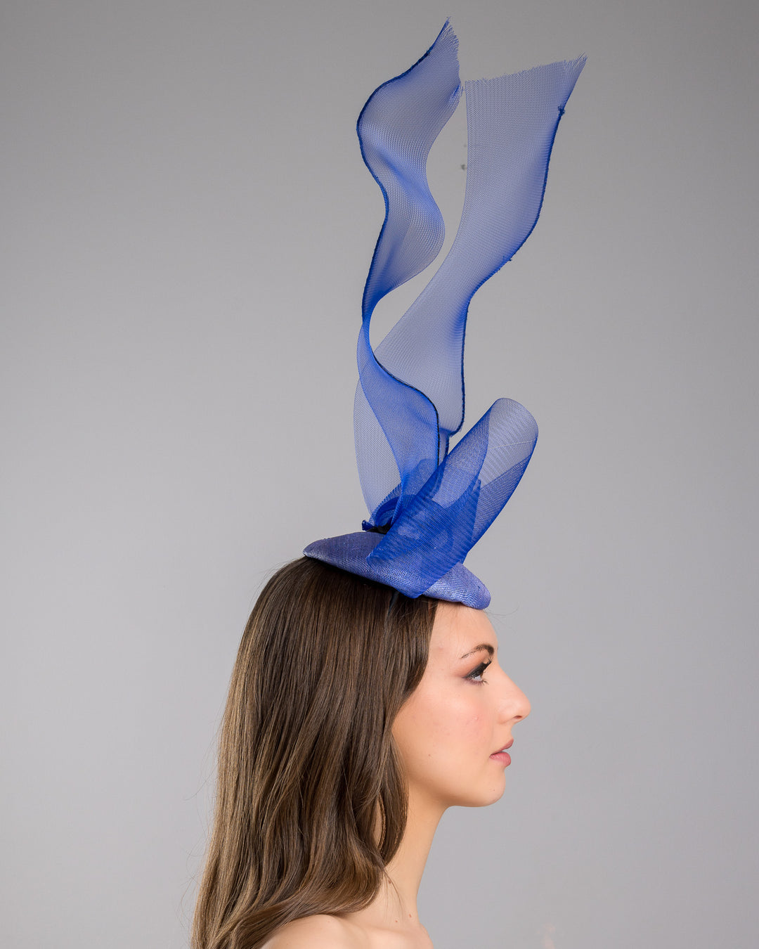 Model is showcasing the right side of the Royal blue button shaped fascinator decorated with a large crinoline bow and flying crinoline ribbons. Made by Merve Bayindir