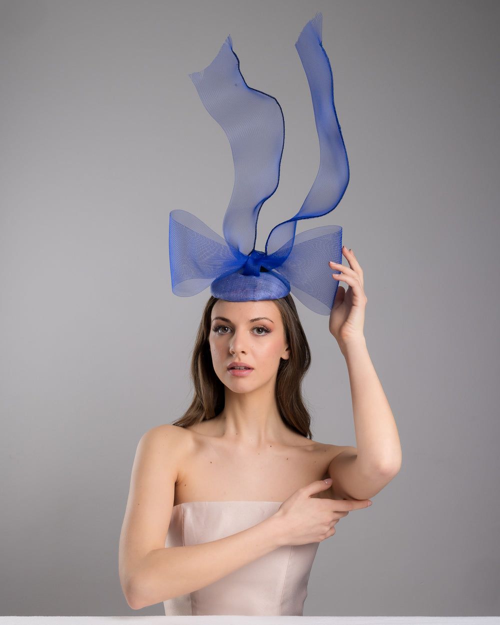 Model is wearing  Royal blue button shaped fascinator decorated with a large crinoline bow and flying crinoline ribbons. Made by Merve Bayindir Millinery.