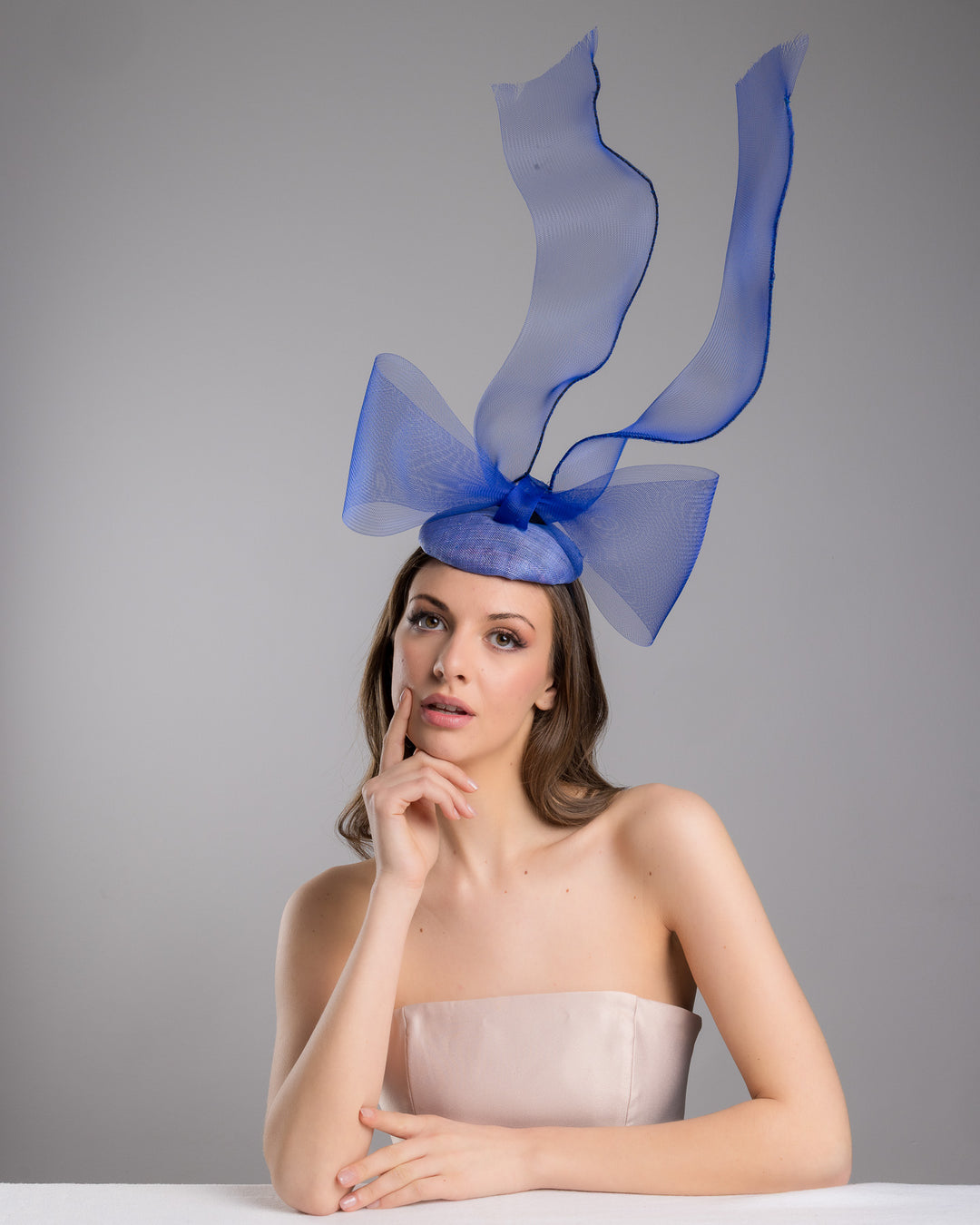 Model is wearing  Royal blue button shaped fascinator decorated with a large crinoline bow and flying crinoline ribbons. Made by Merve Bayindir Millinery.