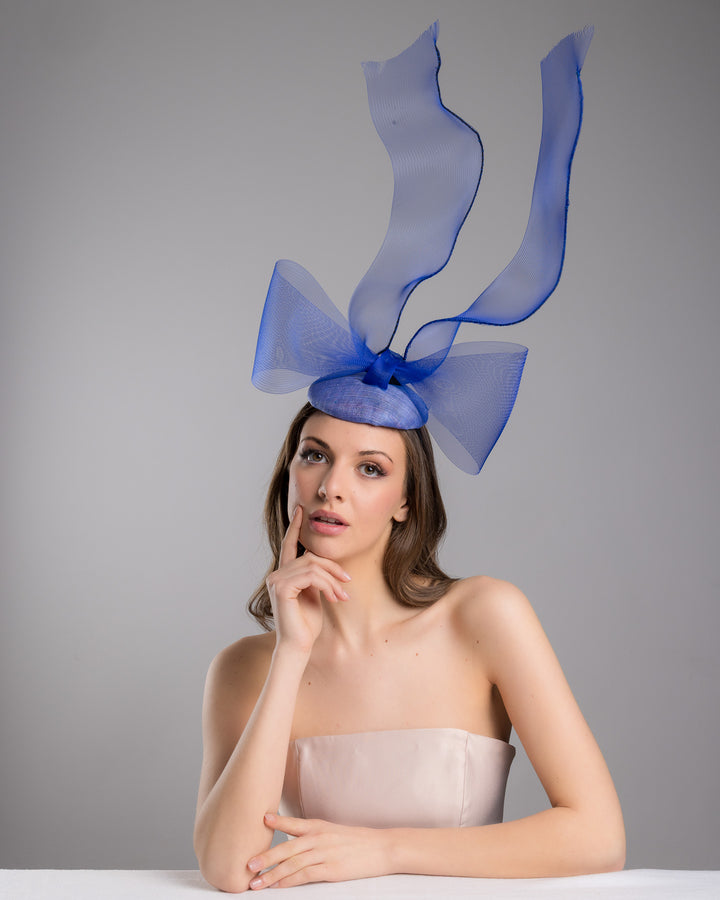 Model is wearing  Royal blue button shaped fascinator decorated with a large crinoline bow and flying crinoline ribbons. Made by Merve Bayindir Millinery.