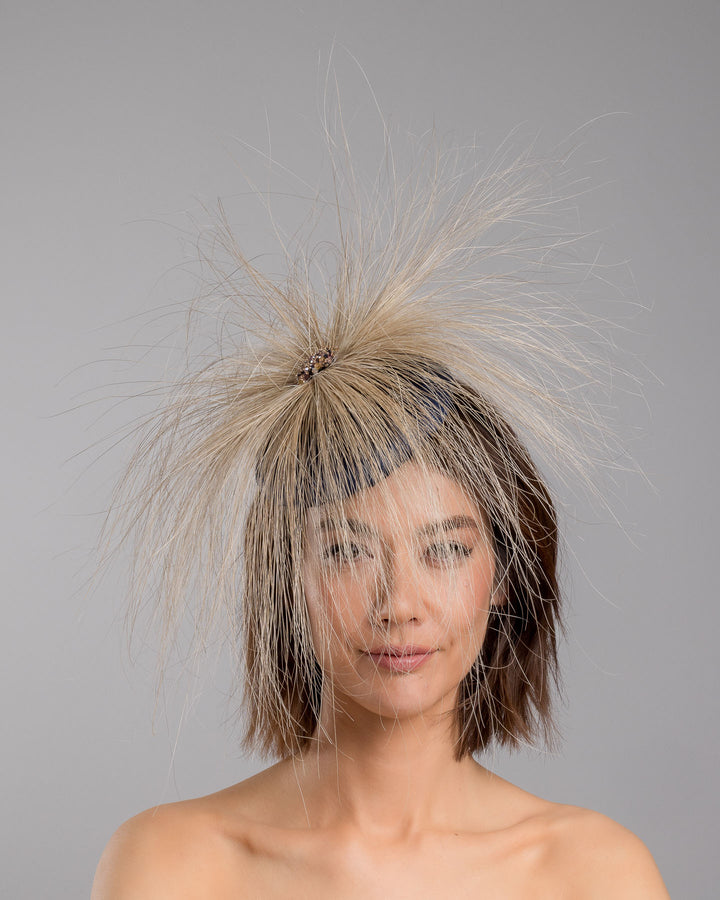 Model is showcasing the front of Navy coloured button shaped fascinator decorated with natural coloured burnt extra long peacock feathers and cluster of crystals. Made by Merve Baindir millinery.