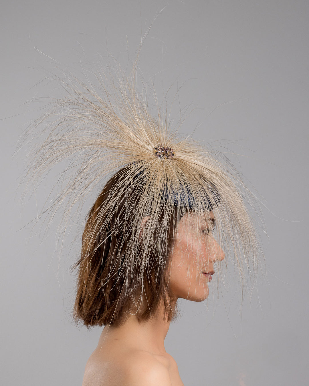 Model is showcasing the right side of Navy coloured button shaped fascinator decorated with natural coloured burnt extra long peacock feathers and cluster of crystals. Made by Merve Baindir millinery.