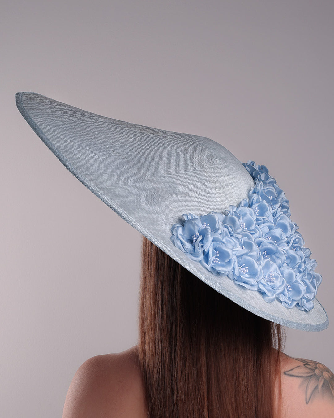 The model presents the back of a blue extra wide brim sinamay hat, tillted to the right side fitted on a mini saucer and secured to the head with wire headband and double elastic band. The right inside of the hat is filled with blue silk flowers and the outer bottom is filled with silked blue flowers creating a visual impact as the flowers from the inner side extending to the outer side.