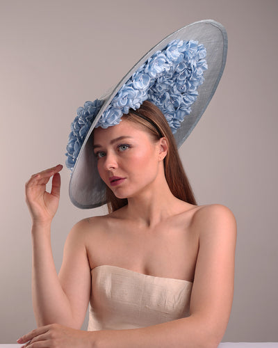 The model presents the left front angle of a blue extra wide brim sinamay hat, tillted to the right side fitted on a mini saucer and secured to the head with wire headband and double elastic band. The right inside of the hat is filled with blue silk flowers and the outer bottom is filled with silked blue flowers creating a visual impact as the flowers from the inner side extending to the outer side.