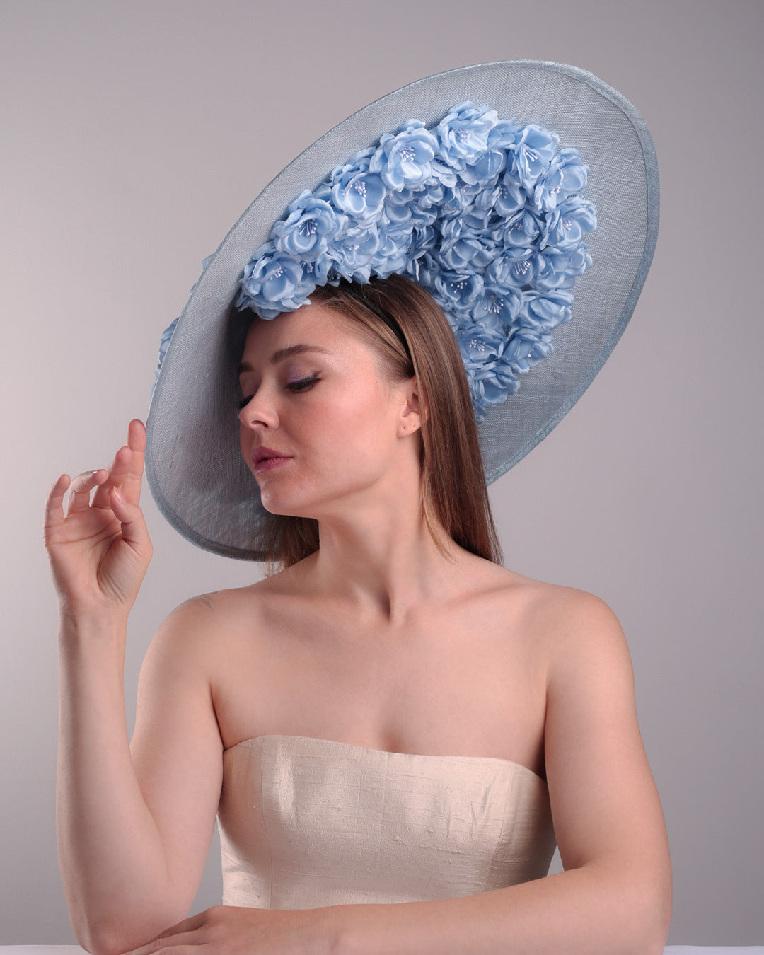 The model presents the right front angle of a blue extra wide brim sinamay hat, tillted to the right side fitted on a mini saucer and secured to the head with wire headband and double elastic band. The right inside of the hat is filled with blue silk flowers and the outer bottom is filled with silked blue flowers creating a visual impact as the flowers from the inner side extending to the outer side.
