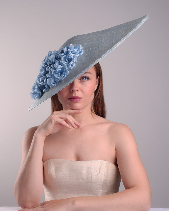 The model presents the front view of a blue extra wide brim sinamay hat, tillted to the right side fitted on a mini saucer and secured to the head with wire headband and double elastic band. The right inside of the hat is filled with blue silk flowers and the outer bottom is filled with silked blue flowers creating a visual impact as the flowers from the inner side extending to the outer side.