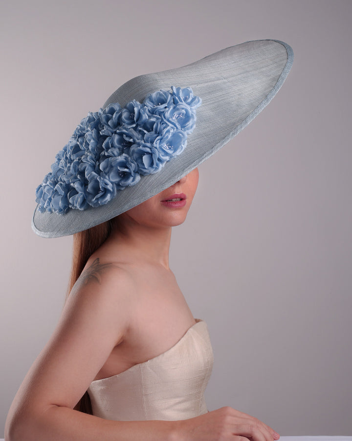The model presents the left front angle from the top of a blue extra wide brim sinamay hat, tillted to the right side fitted on a mini saucer and secured to the head with wire headband and double elastic band. The right inside of the hat is filled with blue silk flowers and the outer bottom is filled with silked blue flowers creating a visual impact as the flowers from the inner side extending to the outer side.