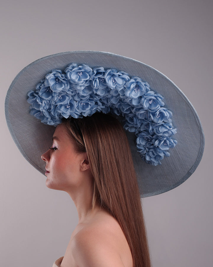The model presents the left side of a blue extra wide brim sinamay hat, tillted to the right side fitted on a mini saucer and secured to the head with wire headband and double elastic band. The right inside of the hat is filled with blue silk flowers and the outer bottom is filled with silked blue flowers creating a visual impact as the flowers from the inner side extending to the outer side.