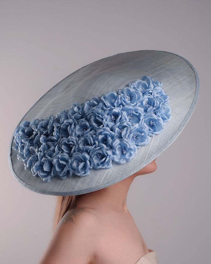 The model presents the right side of a blue extra wide brim sinamay hat, tillted to the right side fitted on a mini saucer and secured to the head with wire headband and double elastic band. The right inside of the hat is filled with blue silk flowers and the outer bottom is filled with silked blue flowers creating a visual impact as the flowers from the inner side extending to the outer side.