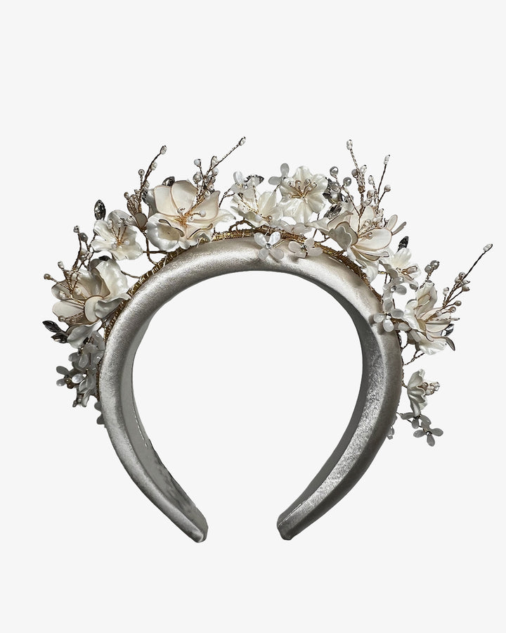 Ivory satin padded headband featuring a crown-like arrangement of handcrafted translucent ivory flowers. The design is accented with fine gold branches, crystal details, pearl-like beads, and subtle grey leaf elements, creating a luminous and romantic statement piece ideal for brides and elegant formal occasions. Designed and made by Merve Bayindir Millinery.

