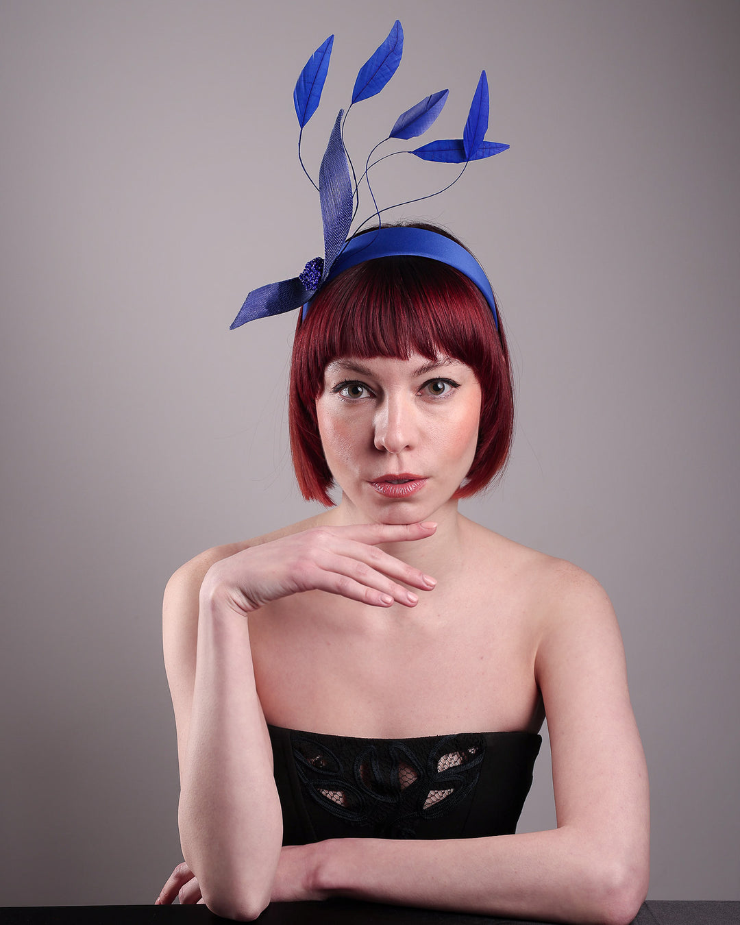 The model showcases the front of the Motmot headband fascinator crafted in a rich cobalt blue. This bold headband features a spray of sculpted feathers with hand-curved edges, accented with a swirl of buntal ribbon tied together with a beaded centre. Designed by Merve Bayindir.