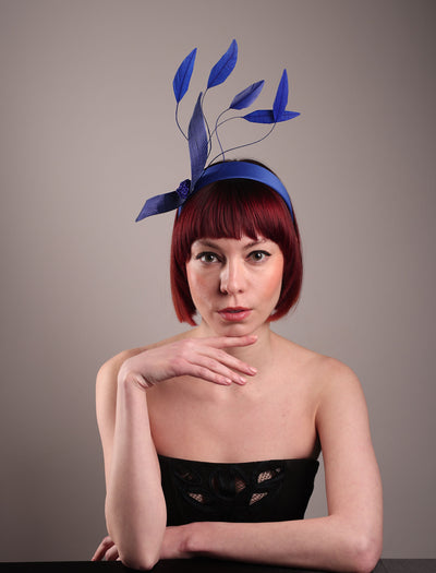 The model showcases the front of the Motmot headband fascinator crafted in a rich cobalt blue. This bold headband features a spray of sculpted feathers with hand-curved edges, accented with a swirl of buntal ribbon tied together with a beaded centre. Designed by Merve Bayindir.