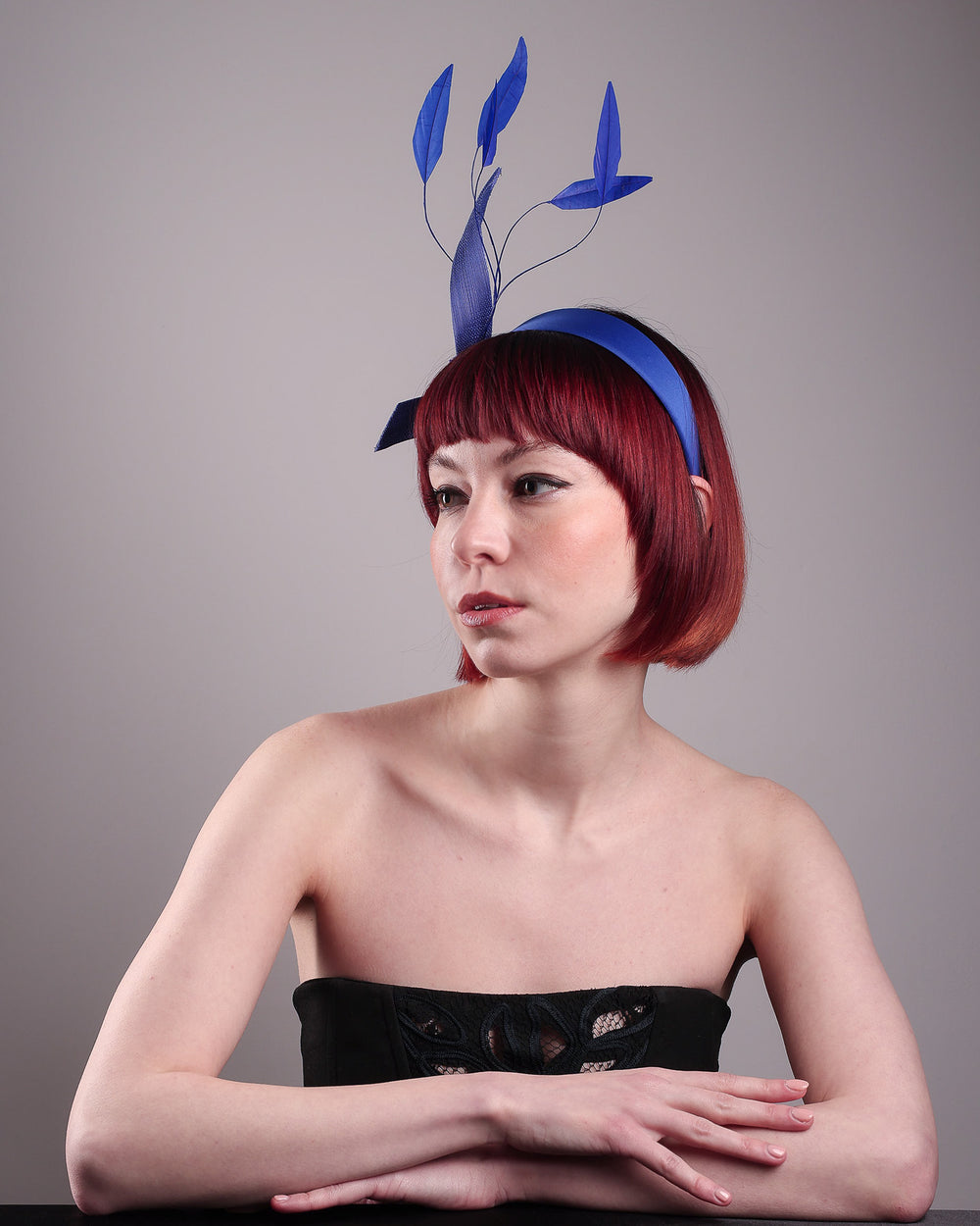 The model showcases the left front of the Motmot headband fascinator crafted in a rich cobalt blue. This bold headband features a spray of sculpted feathers with hand-curved edges, accented with a swirl of buntal ribbon tied together with a beaded centre. Designed by Merve Bayindir.