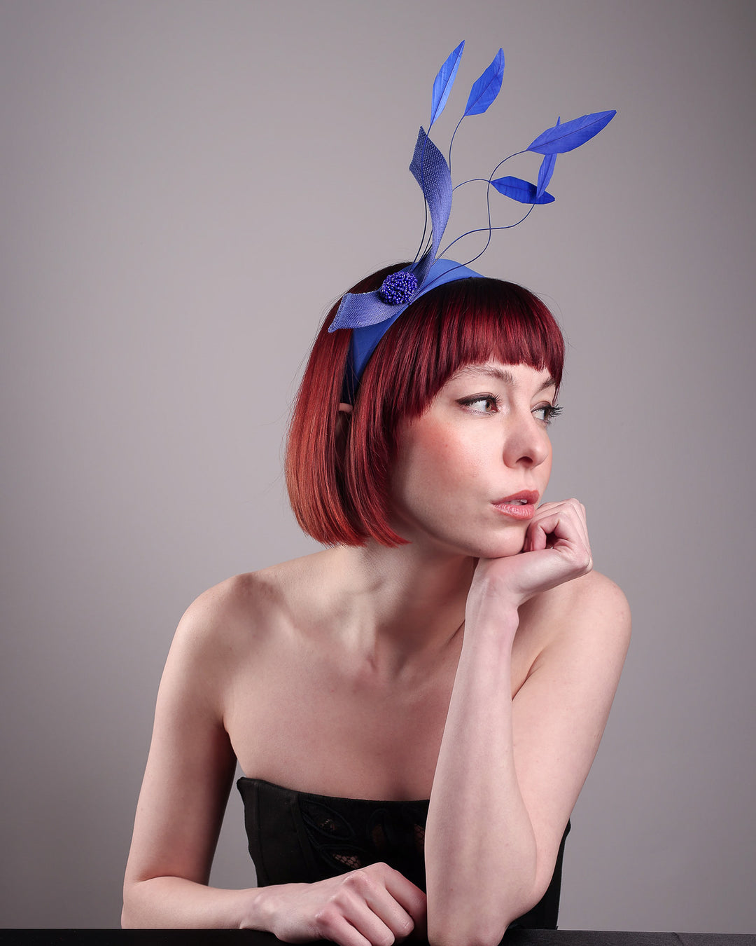 The model showcases the right front of the Motmot headband fascinator crafted in a rich cobalt blue. This bold headband features a spray of sculpted feathers with hand-curved edges, accented with a swirl of buntal ribbon tied together with a beaded centre. Designed by Merve Bayindir.