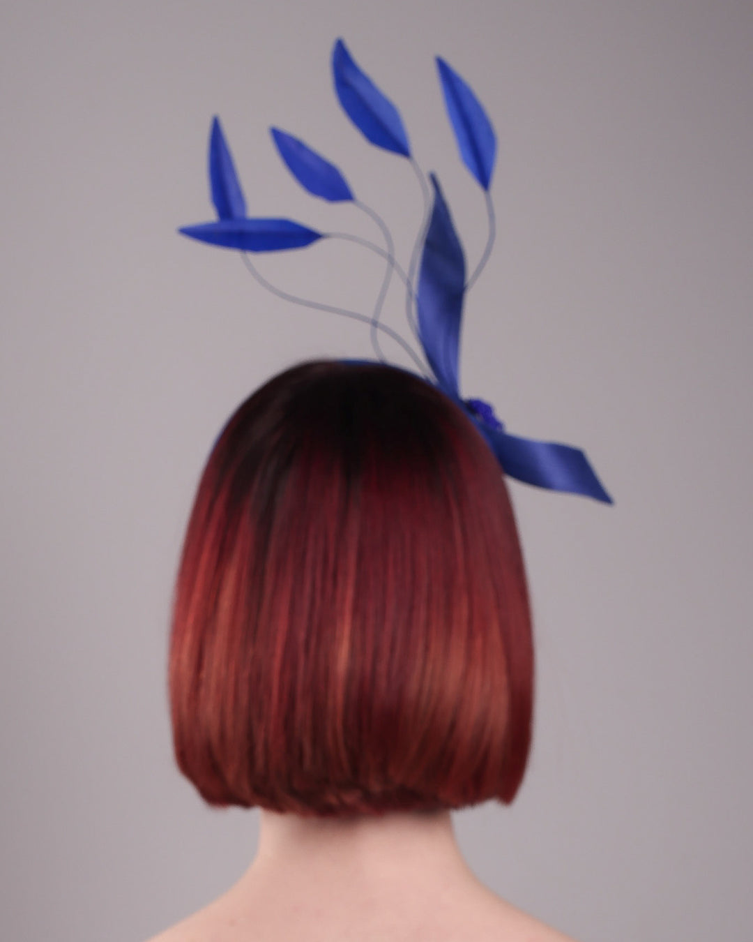 The model showcases the back of the Motmot headband fascinator crafted in a rich cobalt blue. This bold headband features a spray of sculpted feathers with hand-curved edges, accented with a swirl of buntal ribbon tied together with a beaded centre. Designed by Merve Bayindir.