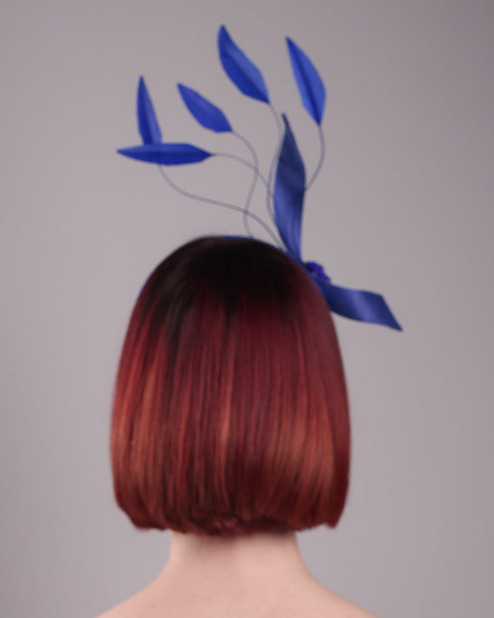 The model showcases the back of the Motmot headband fascinator crafted in a rich cobalt blue. This bold headband features a spray of sculpted feathers with hand-curved edges, accented with a swirl of buntal ribbon tied together with a beaded centre. Designed by Merve Bayindir.