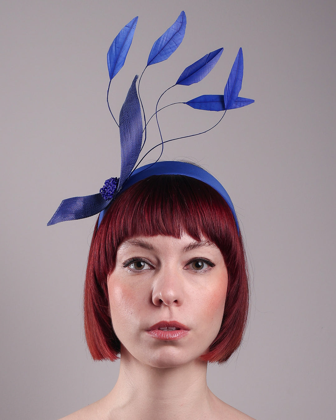 The model showcases the front close up of the Motmot headband fascinator crafted in a rich cobalt blue. This bold headband features a spray of sculpted feathers with hand-curved edges, accented with a swirl of buntal ribbon tied together with a beaded centre. Designed by Merve Bayindir.
