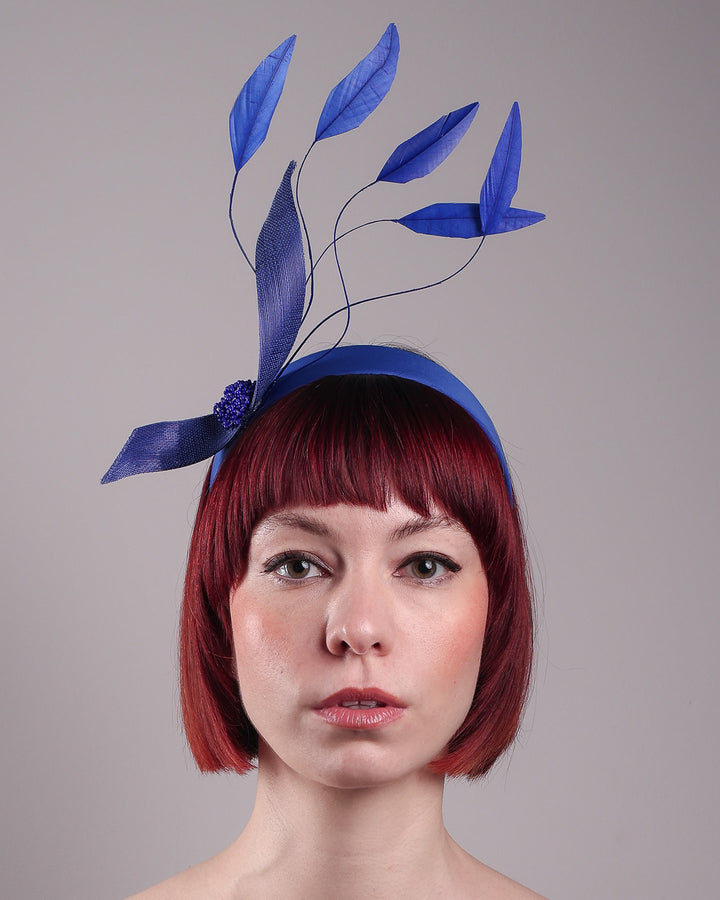 The model showcases the front close up of the Motmot headband fascinator crafted in a rich cobalt blue. This bold headband features a spray of sculpted feathers with hand-curved edges, accented with a swirl of buntal ribbon tied together with a beaded centre. Designed by Merve Bayindir.