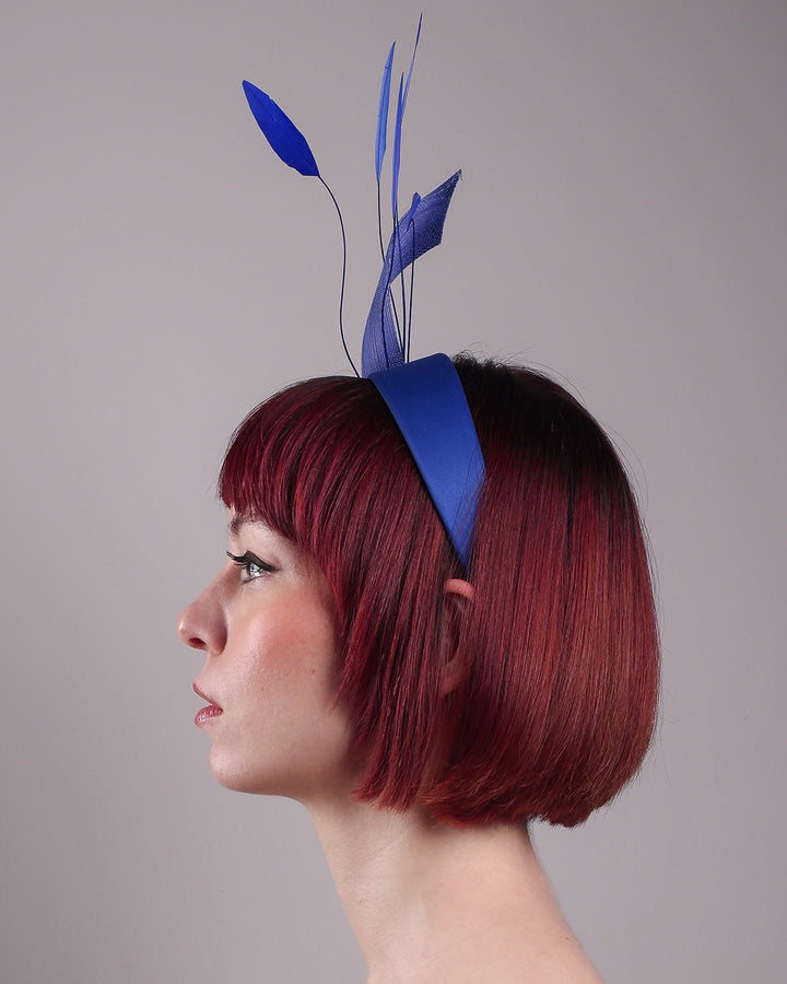 The model showcases the right side close up of the Motmot headband fascinator crafted in a rich cobalt blue. This bold headband features a spray of sculpted feathers with hand-curved edges, accented with a swirl of buntal ribbon tied together with a beaded centre. Designed by Merve Bayindir.